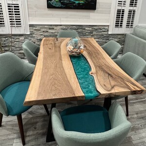 May include: A rectangular dining table featuring a natural live-edge wood slab with a central translucent teal resin river inlay, supported by black metal legs, surrounded by six upholstered chairs with light green backs and teal seats.