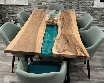 May include: A rectangular live-edge wood dining table featuring a central river inlay made of translucent teal resin. The table is supported by black metal legs and is surrounded by six upholstered chairs in a muted sage green fabric.