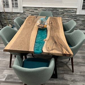 May include: A rectangular live-edge wood dining table featuring a central river inlay made of translucent teal resin. The table is supported by black metal legs and is surrounded by six upholstered chairs in a muted sage green fabric.