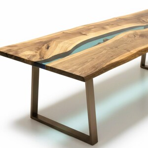 Puede incluir: Una mesa de comedor rectangular con dos tablones de madera de borde natural unidos por un r&iacute;o central de resina azul claro transl&uacute;cida. La mesa se apoya sobre dos patas met&aacute;licas en forma de U con acabado en bronce mate.