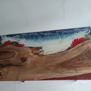 Op de afbeelding: Een rechthoekige tafel met een natuurlijk houten blad met een levende rand, gecombineerd met blauwe en witte hars die oceaangolven nabootst. Het ontwerp bevat ingebedde rode bloemaccenten, kleine schelpjes en een dieprode harsrand langs de zijkant.