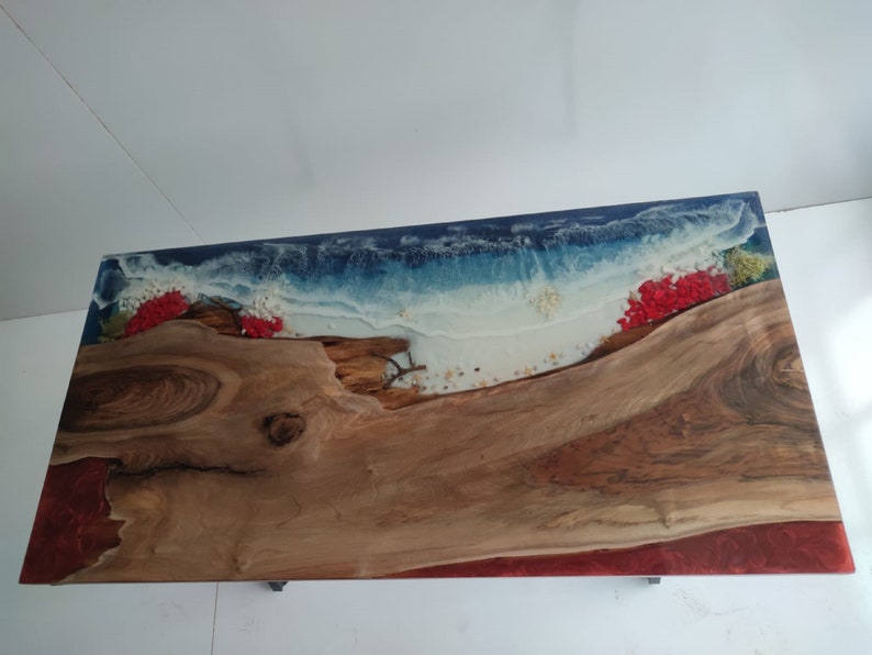 Op de afbeelding: Een rechthoekige tafel met een natuurlijk houten blad met een levende rand, gecombineerd met blauwe en witte hars die oceaangolven nabootst. Het ontwerp bevat ingebedde rode bloemaccenten, kleine schelpjes en een dieprode harsrand langs de zijkant.