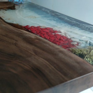 Op de afbeelding: Een houten plank met een natuurlijke rand en een harsinleg die een kustsc&egrave;ne nabootst. Het ontwerp bevat blauw water, wit schuim, rode stenen en groen mos ingebed in de heldere hars, wat contrasteert met de donkerbruine houtnerf.