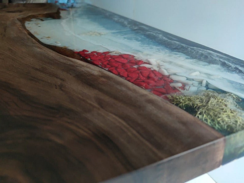 Op de afbeelding: Een houten plank met een natuurlijke rand en een harsinleg die een kustsc&egrave;ne nabootst. Het ontwerp bevat blauw water, wit schuim, rode stenen en groen mos ingebed in de heldere hars, wat contrasteert met de donkerbruine houtnerf.