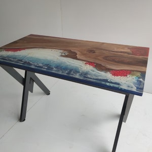 Op de afbeelding: Een rechthoekige tafel met een natuurlijke houten rand gecombineerd met blauwe en witte hars die oceaangolven nabootst. Het oppervlak bevat ingebedde rode decoratieve elementen en wordt ondersteund door zwarte metalen poten met een hoekig ontwerp.