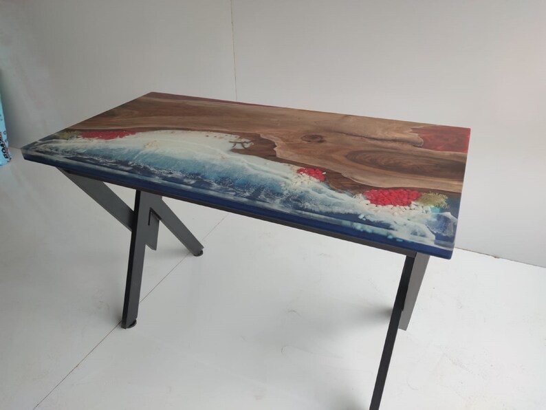 Op de afbeelding: Een rechthoekige tafel met een natuurlijke houten rand gecombineerd met blauwe en witte hars die oceaangolven nabootst. Het oppervlak bevat ingebedde rode decoratieve elementen en wordt ondersteund door zwarte metalen poten met een hoekig ontwerp.