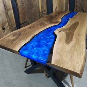 Pu&ograve; includere: Un tavolo in legno con un fiume in resina epossidica blu che attraversa il centro. Il tavolo ha una finitura in legno naturale ed &egrave; sostenuto da una base in metallo nero.