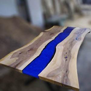 Pu&ograve; includere: Un piano da tavolo in legno con un fiume di resina blu che attraversa il centro. Il legno ha una venatura naturale e la resina &egrave; liscia e lucida.