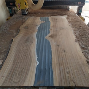 Puede incluir: Una gran losa de madera sin terminar con un borde natural y una incrustaci&oacute;n central de resina epoxi gris-azul. La veta de la madera es visible y la pieza est&aacute; rodeada de virutas de madera, lo que sugiere que est&aacute; en proceso de elaboraci&oacute;n.