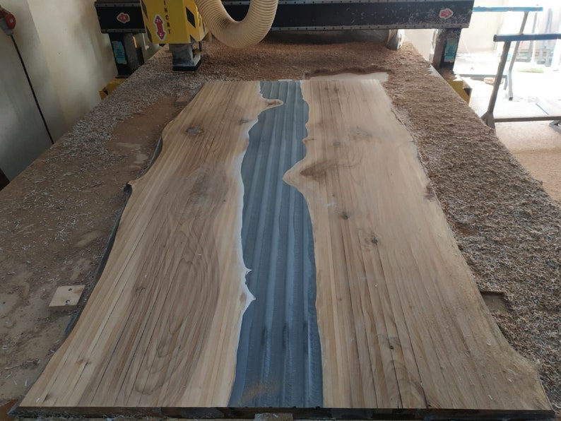 Puede incluir: Una gran losa de madera sin terminar con un borde natural y una incrustaci&oacute;n central de resina epoxi gris-azul. La veta de la madera es visible y la pieza est&aacute; rodeada de virutas de madera, lo que sugiere que est&aacute; en proceso de elaboraci&oacute;n.