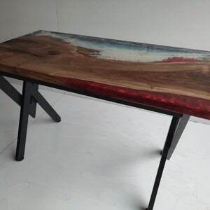 Op de afbeelding: Een rechthoekige tafel met een natuurlijk houten blad met een boomkant, ge&iuml;ntegreerd met doorschijnende blauwe en rode harsaccenten die een kustlijn nabootsen. De tafel wordt ondersteund door een zwart metalen frame met schuine poten en een kruisverbinding.