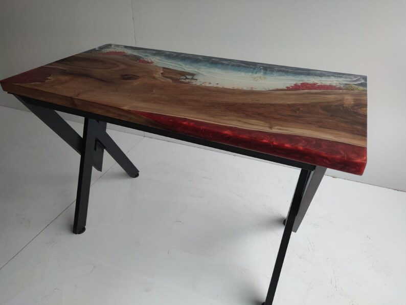 Op de afbeelding: Een rechthoekige tafel met een natuurlijk houten blad met een boomkant, ge&iuml;ntegreerd met doorschijnende blauwe en rode harsaccenten die een kustlijn nabootsen. De tafel wordt ondersteund door een zwart metalen frame met schuine poten en een kruisverbinding.