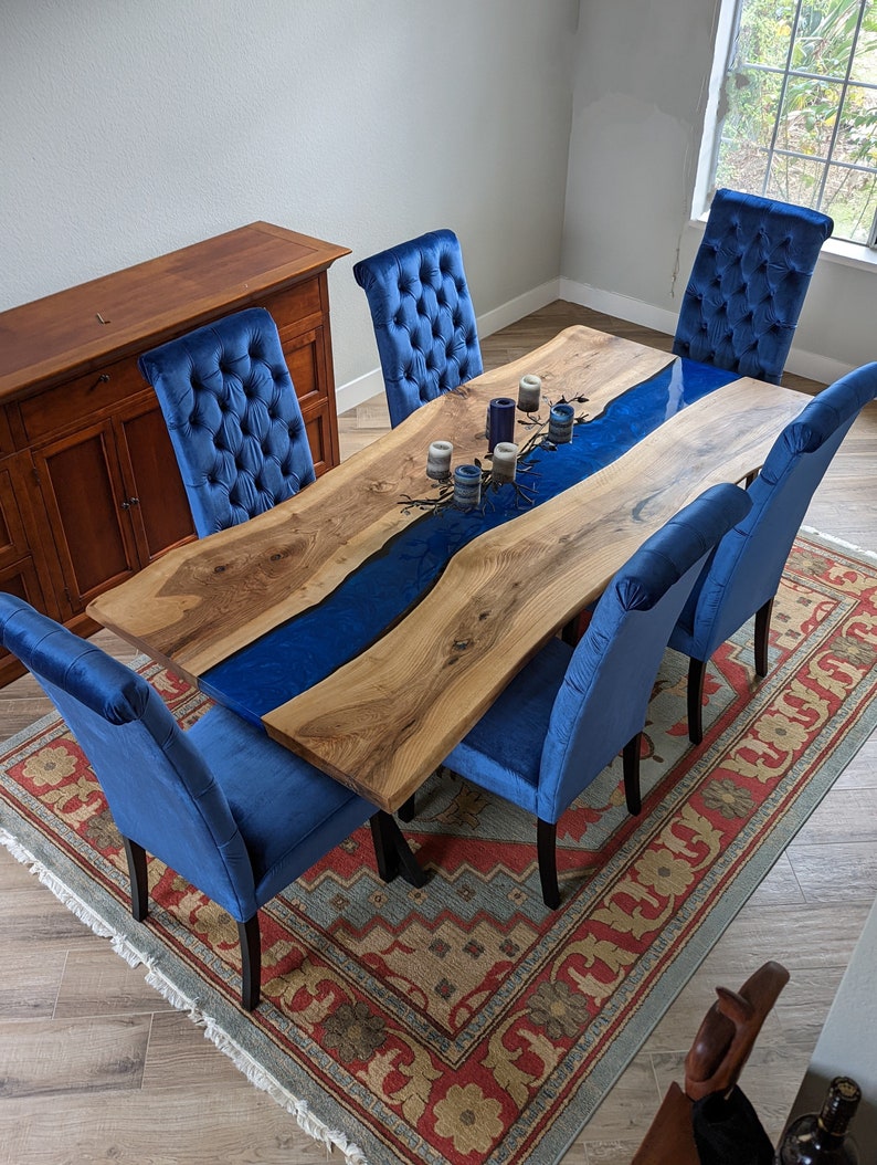 Pu&ograve; includere: Un tavolo in legno naturale con un fiume in resina blu e sei sedie in velluto blu. Il tavolo &egrave; apparecchiato con candele ed &egrave; circondato da un tappeto con un motivo floreale.