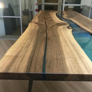 Puede incluir: Una mesa de comedor de madera con borde natural y una incrustaci&oacute;n central de resina azul transl&uacute;cida. La veta de la madera muestra tonos marrones claros y oscuros con nudos visibles, creando un efecto similar a un r&iacute;o.