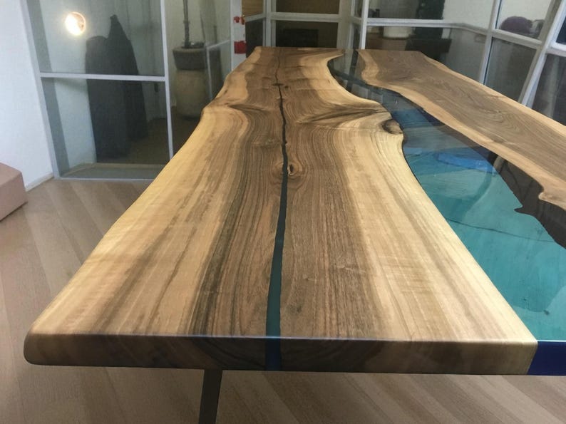 Puede incluir: Una mesa de comedor de madera con borde natural y una incrustaci&oacute;n central de resina azul transl&uacute;cida. La veta de la madera muestra tonos marrones claros y oscuros con nudos visibles, creando un efecto similar a un r&iacute;o.