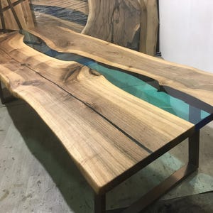 Puede incluir: Una mesa de comedor rectangular con dos tablones de madera de nogal de borde natural unidos por una incrustaci&oacute;n de resina transl&uacute;cida de color verde azulado. La mesa se apoya sobre una base de metal oscuro.