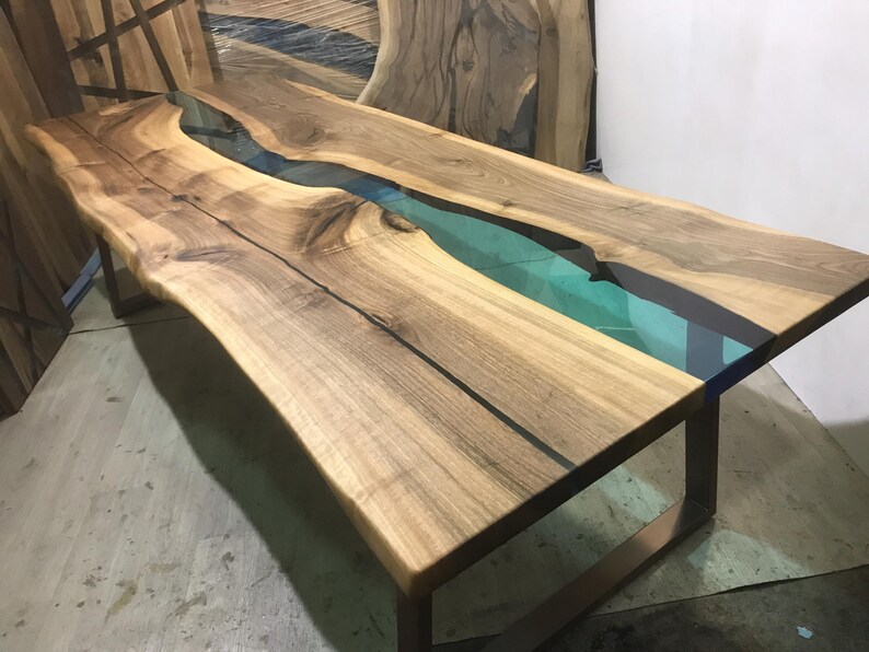 Puede incluir: Una mesa de comedor rectangular con dos tablones de madera de nogal de borde natural unidos por una incrustaci&oacute;n de resina transl&uacute;cida de color verde azulado. La mesa se apoya sobre una base de metal oscuro.