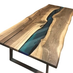 Puede incluir: Una mesa de comedor rectangular con un tablero de madera de borde natural y un patr&oacute;n de veta prominente. Un r&iacute;o de resina azul transl&uacute;cida recorre el centro de la madera. La mesa se apoya sobre una base de metal oscuro en forma de U.