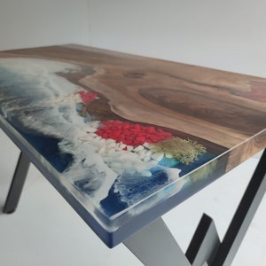 Op de afbeelding: Een rechthoekige tafel met een houten blad met natuurlijke rand, verbonden met blauwe hars. Het harsgedeelte bevat witte schuimeffecten, rode en witte decoratieve stenen en mosachtige texturen. De tafel wordt ondersteund door schuine zwarte metalen poten.