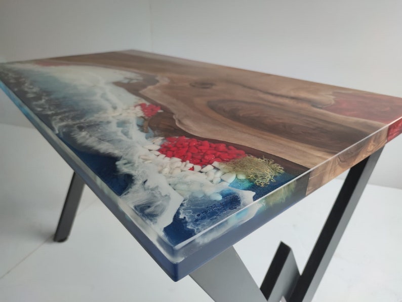 Op de afbeelding: Een rechthoekige tafel met een houten blad met natuurlijke rand, verbonden met blauwe hars. Het harsgedeelte bevat witte schuimeffecten, rode en witte decoratieve stenen en mosachtige texturen. De tafel wordt ondersteund door schuine zwarte metalen poten.
