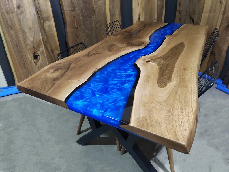 Pu&ograve; includere: Un tavolo in legno con un fiume in resina epossidica blu che attraversa il centro. Il tavolo ha una finitura in legno naturale ed &egrave; sostenuto da una base in metallo nero.
