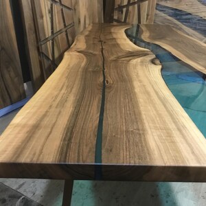 Puede incluir: Una mesa de madera con borde natural que presenta un dise&ntilde;o de r&iacute;o central hecho de resina transl&uacute;cida de color verde azulado. El tablero muestra patrones de veta de madera natural y un acabado liso, con la secci&oacute;n de resina a lo largo.