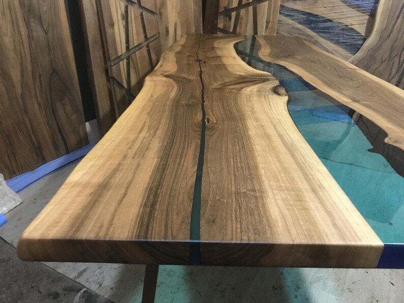 Puede incluir: Una mesa de madera con borde natural que presenta un dise&ntilde;o de r&iacute;o central hecho de resina transl&uacute;cida de color verde azulado. El tablero muestra patrones de veta de madera natural y un acabado liso, con la secci&oacute;n de resina a lo largo.