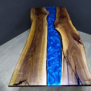 May include: A live edge wood table featuring a central river design made of vibrant blue epoxy resin. The natural wood grain displays light and dark brown tones with visible knots and organic edges, contrasting with the swirling blue resin.