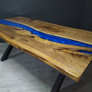 Puede incluir: Una mesa de comedor rectangular con un tablero de madera natural de borde irregular y una incrustación central fluida de resina azul vibrante. La mesa se apoya en patas de metal negro inclinadas y resistentes, que contrastan con la veta orgánica de la madera.
