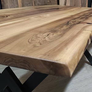 May include: A large, live edge wooden table top with a natural wood finish. The table top is supported by a black metal X-shaped base.