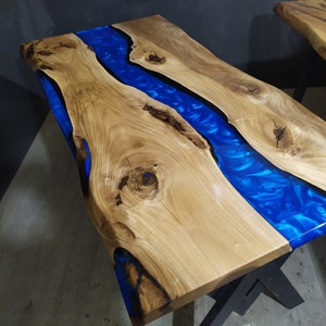 Pu&ograve; includere: Un piano da tavolo in legno con un fiume di resina blu che attraversa il centro. Il legno &egrave; di colore marrone chiaro e la resina &egrave; di un blu intenso. Il piano da tavolo &egrave; su una base in metallo nero.