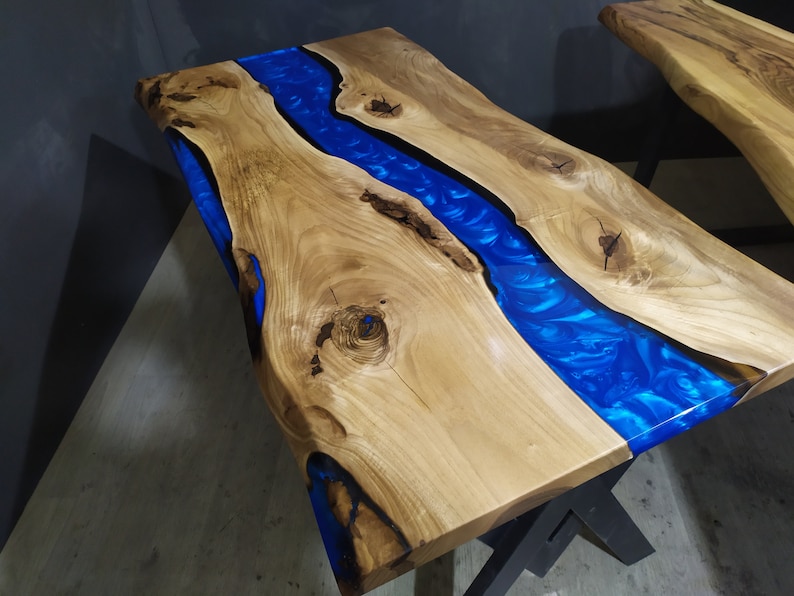 Pu&ograve; includere: Un piano da tavolo in legno con un fiume di resina blu che attraversa il centro. Il legno &egrave; di colore marrone chiaro e la resina &egrave; di un blu intenso. Il piano da tavolo &egrave; su una base in metallo nero.