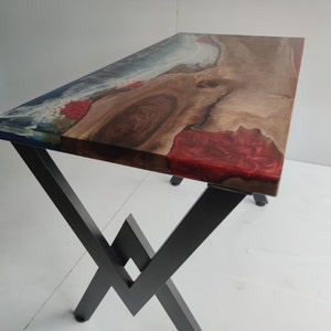 Op de afbeelding: Een rechthoekige tafel met een natuurlijk houten blad met een boomkant, ge&iuml;ntegreerd met secties van doorschijnende blauwe en ondoorzichtige rode hars. Het tafelblad rust op een zwart metalen onderstel met een hoekig X-vormig ontwerp.