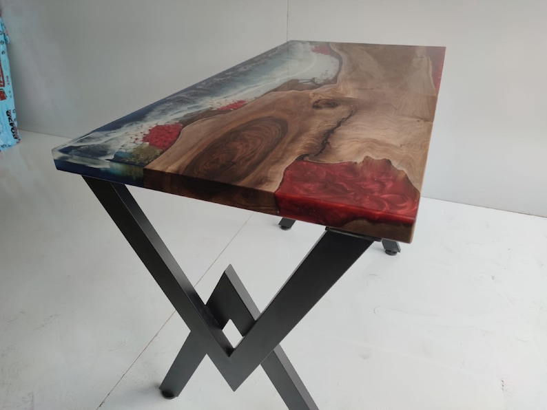 Op de afbeelding: Een rechthoekige tafel met een natuurlijk houten blad met een boomkant, ge&iuml;ntegreerd met secties van doorschijnende blauwe en ondoorzichtige rode hars. Het tafelblad rust op een zwart metalen onderstel met een hoekig X-vormig ontwerp.