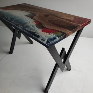 Op de afbeelding: Een rechthoekige tafel met een natuurlijk houten blad met een boomkant, ge&iuml;ntegreerd met blauwe en witte hars die oceaangolven en rode accenten nabootst. De tafel wordt ondersteund door een zwart metalen frame met een geometrisch pootontwerp.