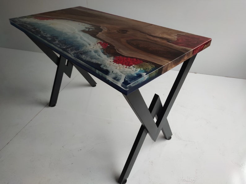 Op de afbeelding: Een rechthoekige tafel met een natuurlijk houten blad met een boomkant, ge&iuml;ntegreerd met blauwe en witte hars die oceaangolven en rode accenten nabootst. De tafel wordt ondersteund door een zwart metalen frame met een geometrisch pootontwerp.