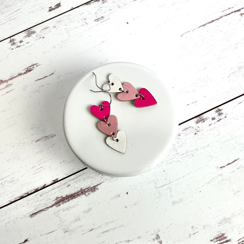 May include: A pair of dangle earrings with three layered hearts. The hearts are pink, white, and a light pink color. The earrings are on a white surface.