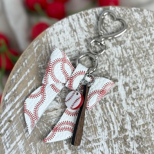 May include: A baseball-themed keychain featuring a white bow with red stitching, a baseball charm, and a wooden bat charm. The keychain has a silver heart-shaped clasp.