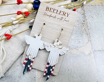 May include: White eagle-shaped earrings with gold-colored hoops and fringed tails featuring a red, white, and blue American flag design. The earrings are displayed on a wooden tag with the text "BEELERY Handmade with love."