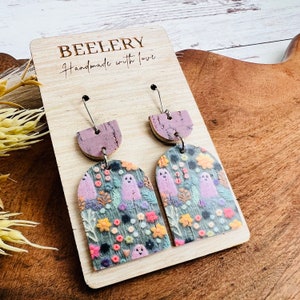 May include: A pair of dangle earrings with a purple and cork base and a green, pink, and white ghost and floral pattern. The earrings are on a wooden display card with the text "BEELERY Handmade with love".