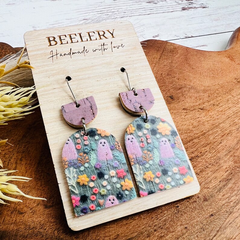 May include: A pair of dangle earrings with a purple and cork base and a green, pink, and white ghost and floral pattern. The earrings are on a wooden display card with the text "BEELERY Handmade with love".