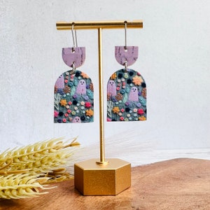 May include: A pair of dangle earrings with a purple and green floral pattern featuring ghosts. The earrings are made of polymer clay and have a cork backing.