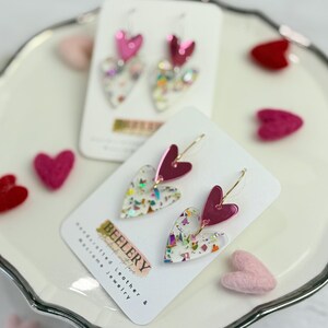 May include: Heart-shaped earrings with a clear base filled with colorful confetti and a magenta heart accent. The earrings are displayed on a white card with the brand name "BEELERY" and are set on a white decorative plate with felt hearts.