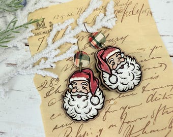 May include: Wooden Santa Claus earrings with red hats and white beards. The earrings have a plaid fabric accent and are hanging on a vintage paper background. Festive Christmas jewelry.