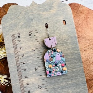 May include: A pair of purple and gray ghost earrings with a floral pattern. The earrings are made of wood and have a cork backing. The earrings are approximately 2 inches long.