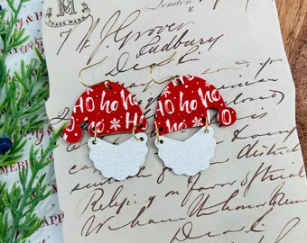 May include: Red and white Santa hat earrings with a white glitter beard. The hat has the words "Ho Ho Ho" printed on it.