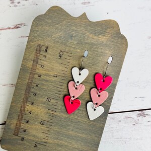 May include: A pair of dangle earrings with three layered hearts. The hearts are pink, white, and a bright pink color. The earrings are on a wooden surface with a ruler.