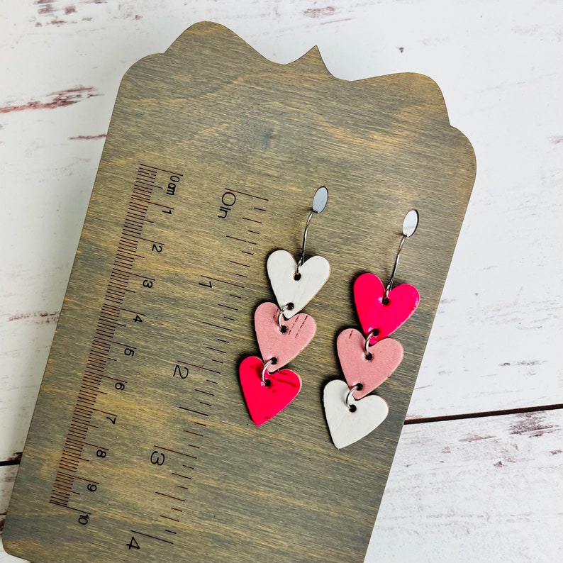 May include: A pair of dangle earrings with three layered hearts. The hearts are pink, white, and a bright pink color. The earrings are on a wooden surface with a ruler.