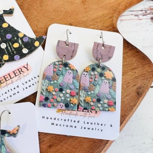 May include: A pair of handmade earrings featuring a ghost and floral pattern on a green background. The earrings are made of leather and have a cork accent. The earrings are handmade with love and are part of a macrame jewelry collection.