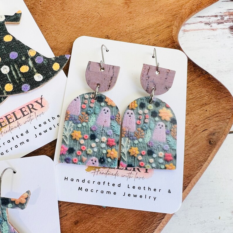 May include: A pair of handmade earrings featuring a ghost and floral pattern on a green background. The earrings are made of leather and have a cork accent. The earrings are handmade with love and are part of a macrame jewelry collection.